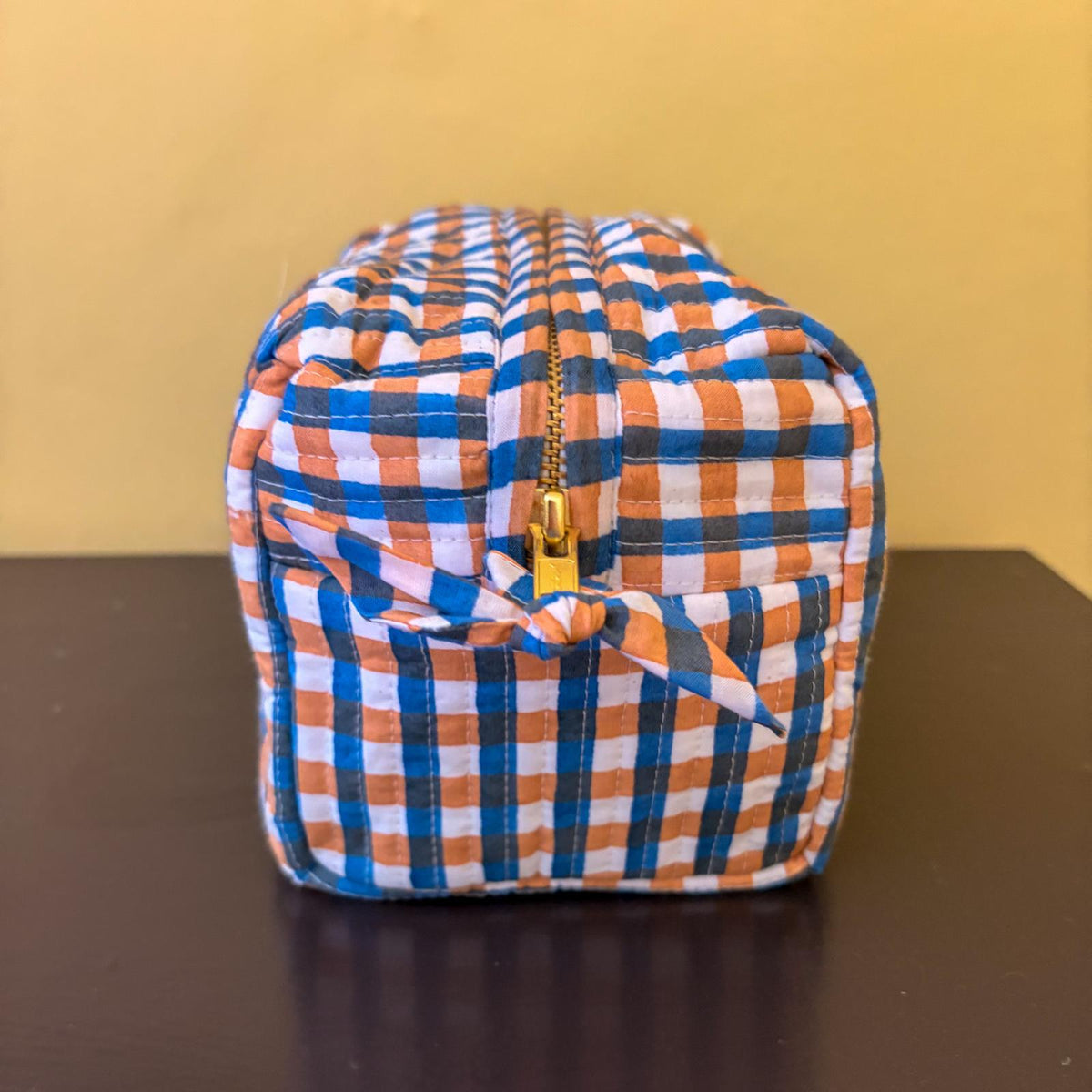 A small zippered pouch with a blue, orange, and white gingham check pattern, featuring a gold zipper and a matching fabric tie bow, set against a yellow background.
