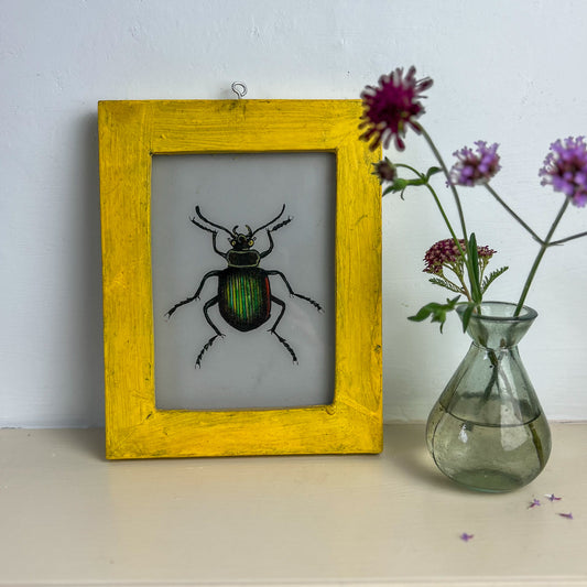 Framed artwork of a beetle on a white wall with a vase of flowers. INDIAN-GLASS-REVERSE-PAINTING-GREEN-BEETLE