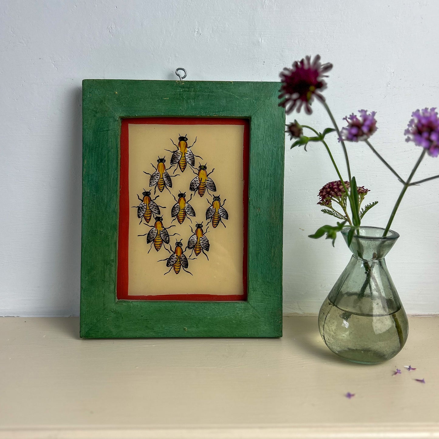 Framed artwork of bees on a beige background with a green frame, next to a vase with flowers on a white surface. INDIAN-GLASS-REVERSE-PAINTING-GROUP-BEES