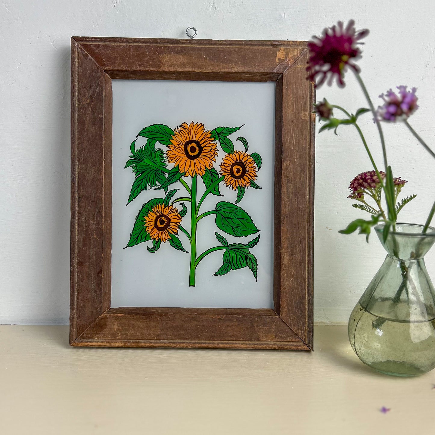 Framed artwork of sunflowers on a white background with a vase of flowers beside it. INDIAN-GLASS-REVERSE-PAINTING-GROUP-SUNFLOWER