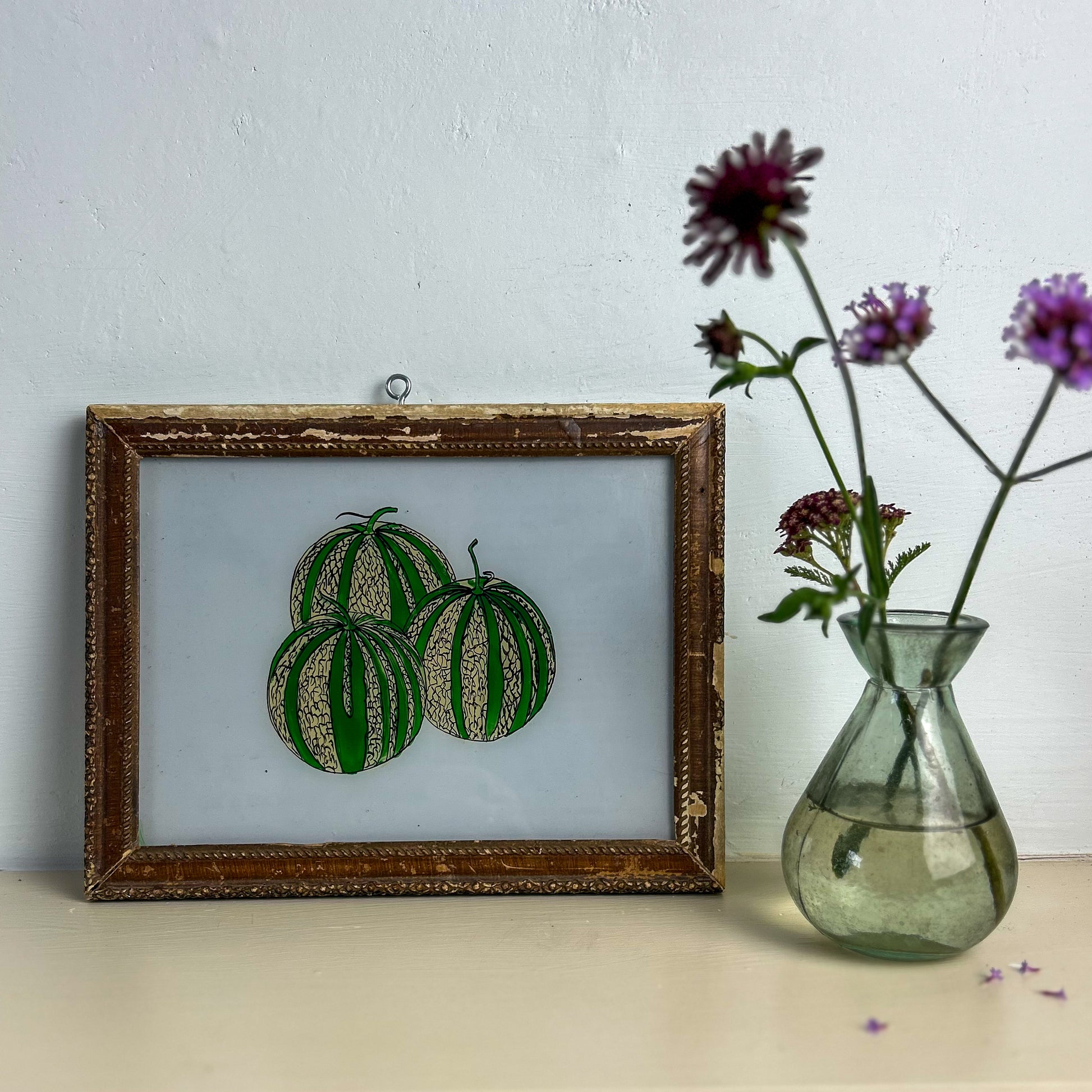 Framed artwork of green fruits on a light blue background next to a vase with flowers.  INDIAN-GLASS-REVERSE-PAINTING-THREE-WATERMELONS