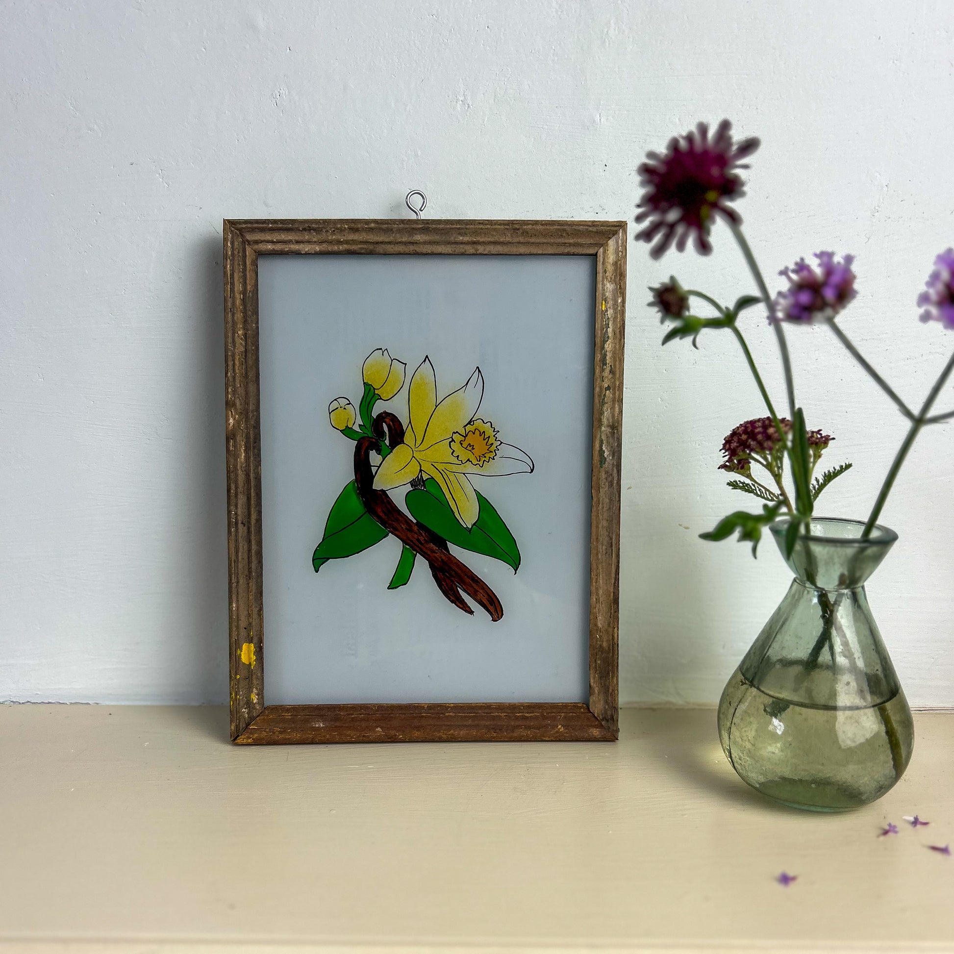 Framed stained glass artwork of a flower on a white wall with a vase of flowers beside it. INDIAN-GLASS-REVERSE-PAINTING-WHITE-FLOWER