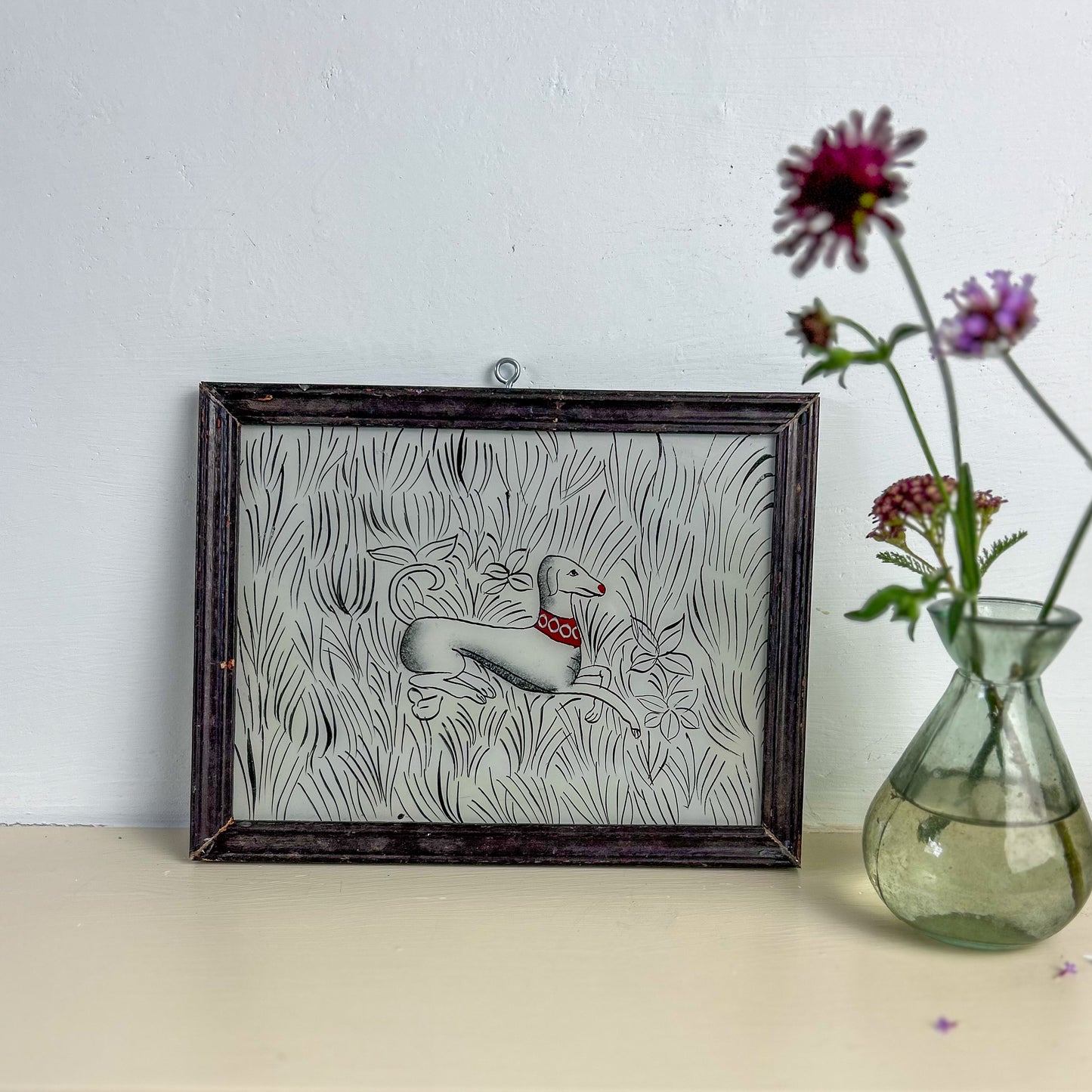 Decorative wall art with a dog design in a wooden frame next to a vase with flowers on a light surface. INDIAN-GLASS-REVERSE-PAINTING-WHITE_DOG
