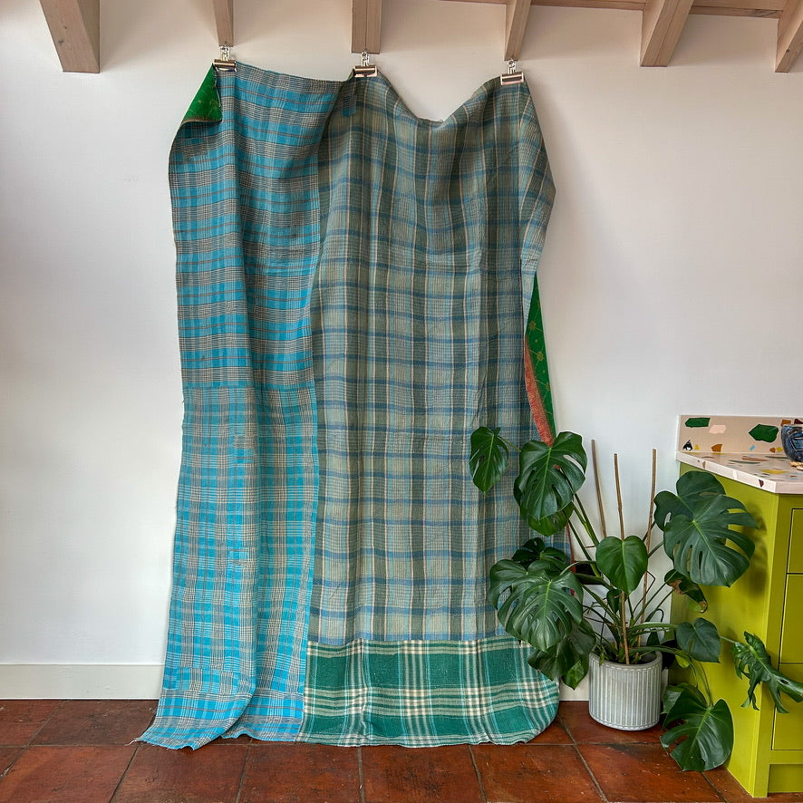 Plaid fabric hanging on a rack against a white wall with a plant and green cabinet in the background.kantha-vintage-2026-hand-made-blanket-throw-textiles-wall-hanging