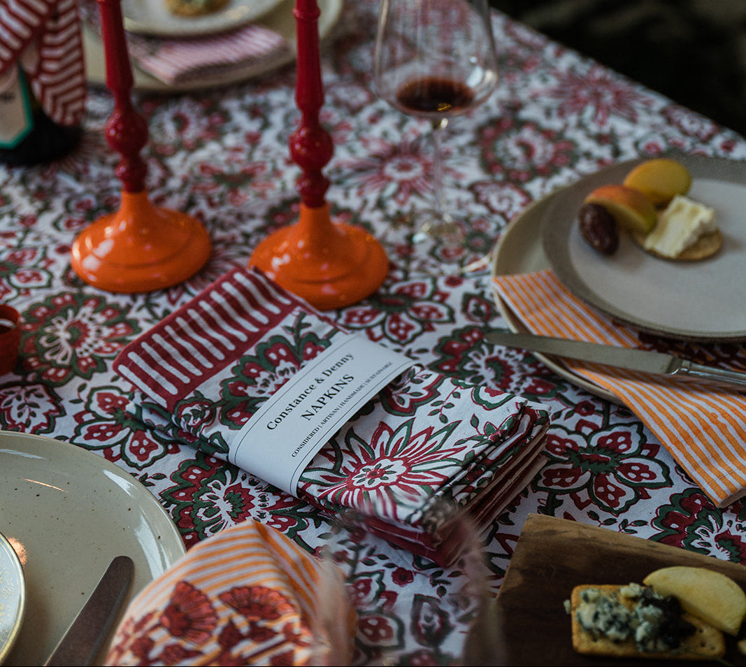 A styled holiday table setting with a red and white patterned tablecloth, two orange taper candles in red holders, plates with cheese and crackers, a wine glass, and a napkin with a 'Gourmet Snacks & Wine' tag, set against a dark festive background with warm string lights.