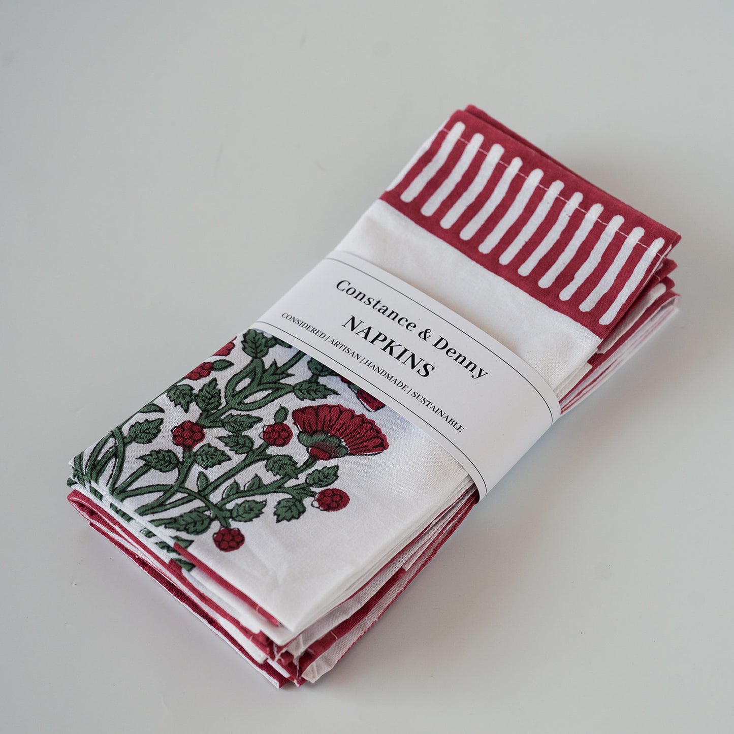 A stack of Constance & Denny napkins in a white paper sleeve, featuring a red and white striped napkin, a red and white checkered napkin, and a white napkin with a red poppy and green leaf pattern.