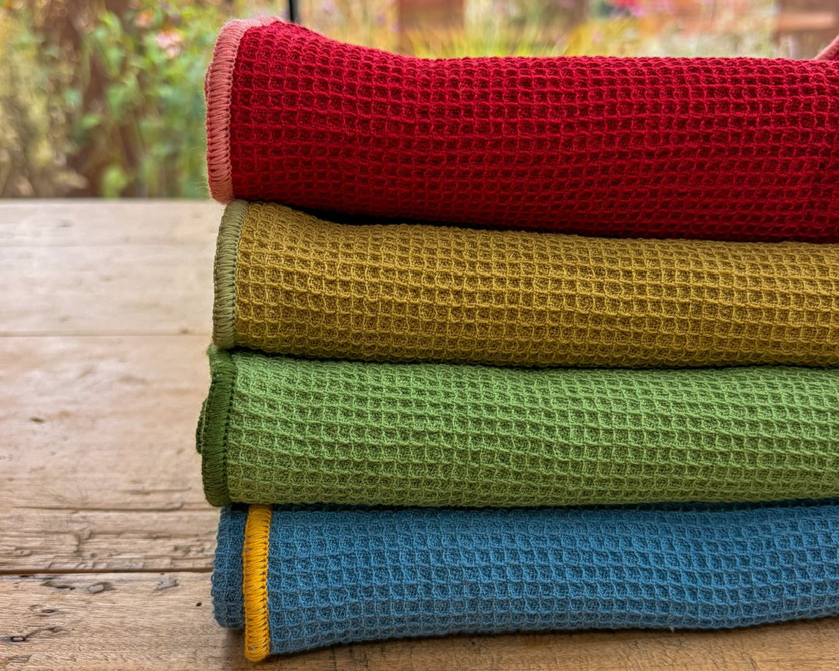 Four folded textured waffle-weave towels stacked on a rustic wooden table, showing red, mustard yellow, green, and blue colors with a thin yellow stripe on the bottom edge.