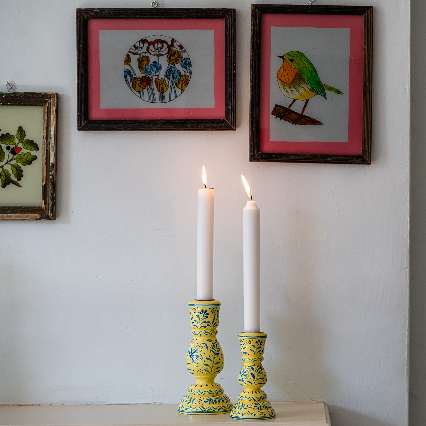 Two lit candles in decorative yellow candlesticks with framed artwork on a wall. constance-and-denny-candle-stick-holder-hand-painted-floral. constance-and-denny-candle-stick-holder-cream-hand-painted-wooden. Brighten your home with this hand-painted ceramic candle holder in yellow & blue. Fits taper candles perfectly. A bohemian, artisan-inspired décor accent for dining tables, mantels & more.