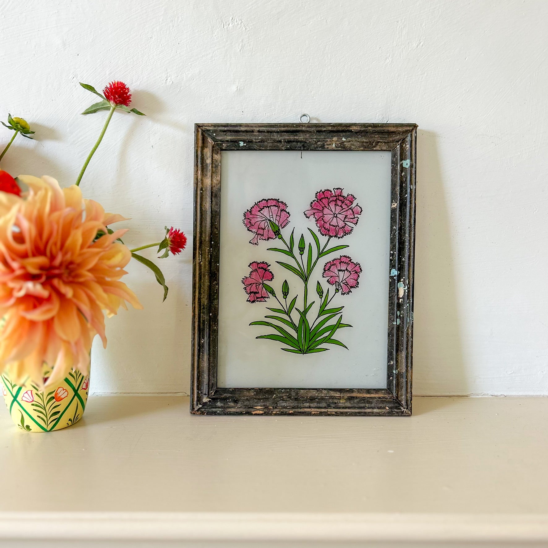 Framed floral artwork with pink flowers on a white surface next to a vase with flowers. constance-and-denny-indian-reverse-glass-painting-boxed-packaging-hand-painted-artisan-art-carnations