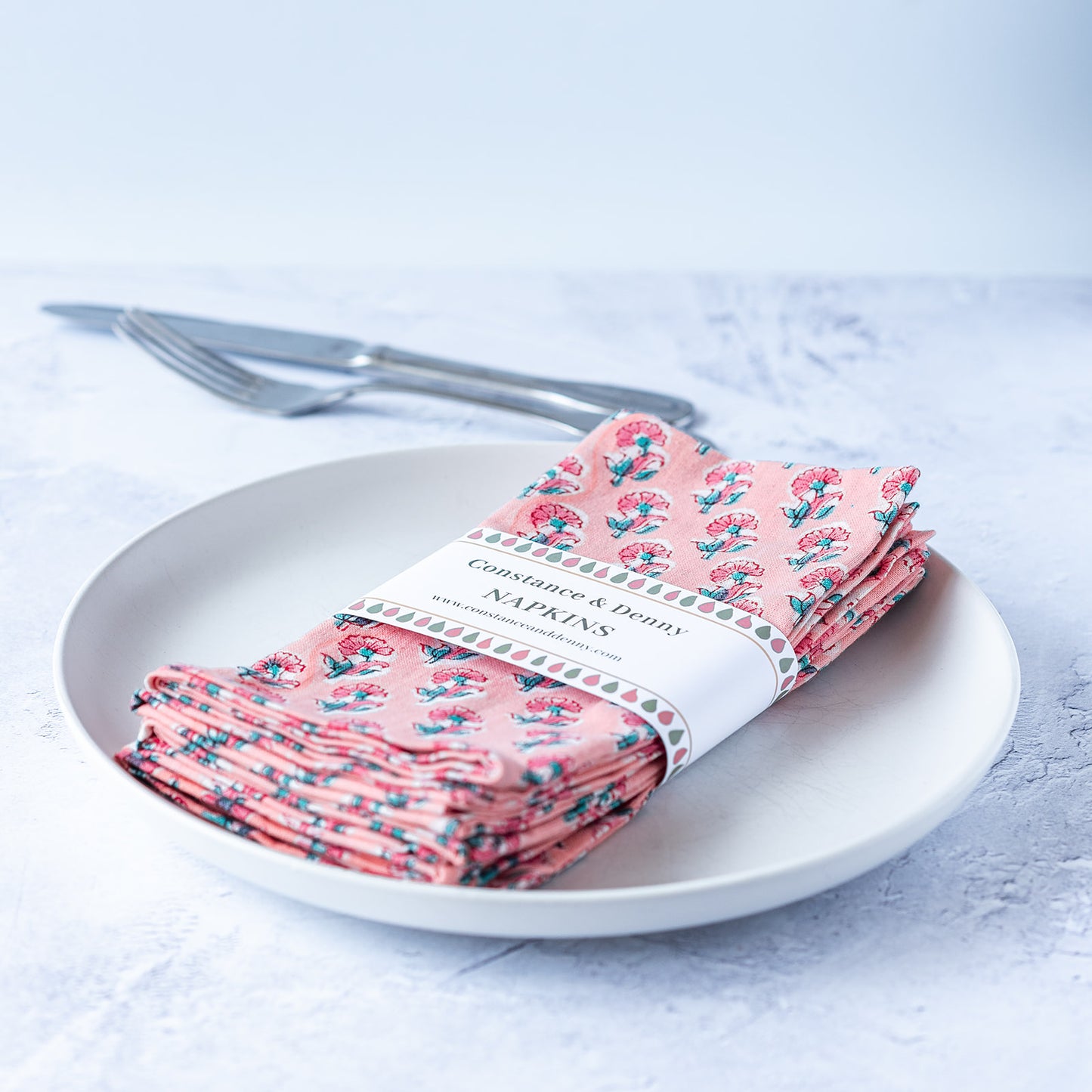 Packaged pink floral napkins on a white plate with cutlery. block-printed-set-of-4-napkins-pink-floral