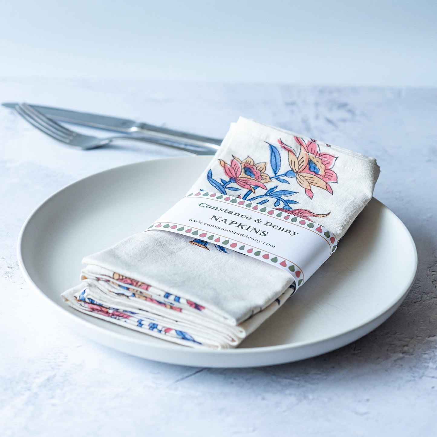Set of floral-patterned napkins on a plate with cutlery against a light background. FLORAL-GREEN-INDIGO-STEM
