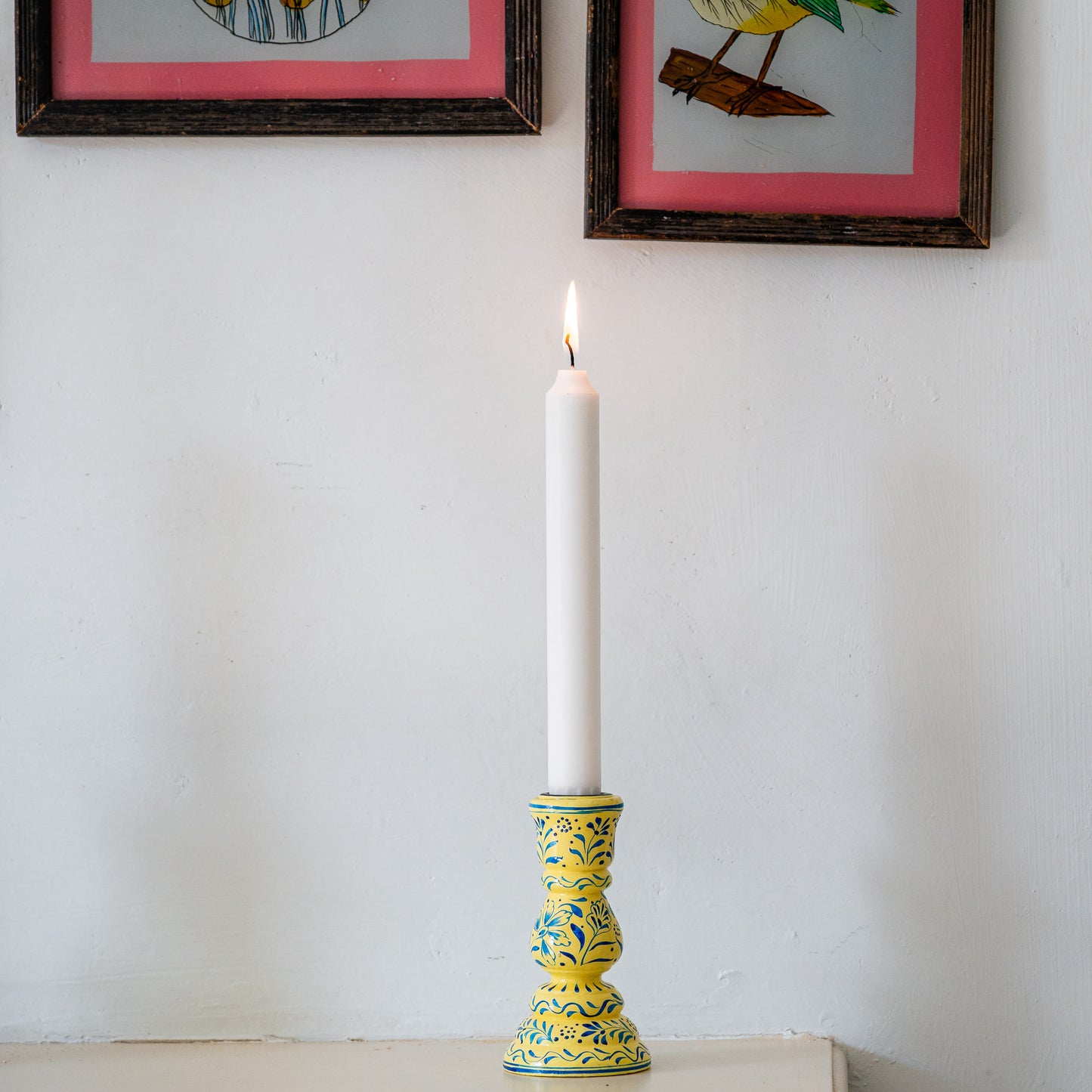 Decorative candle holder with a lit candle against a white wall with framed pictures. constance-and-denny-candle-stick-holder-cream-hand-painted-wooden