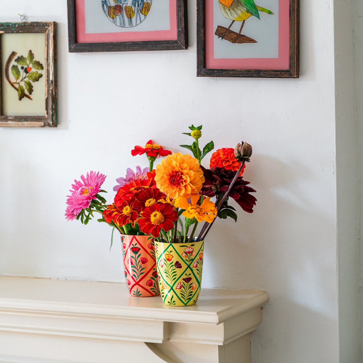 Colorful flowers in patterned vases on a white surface with framed artwork in the background.