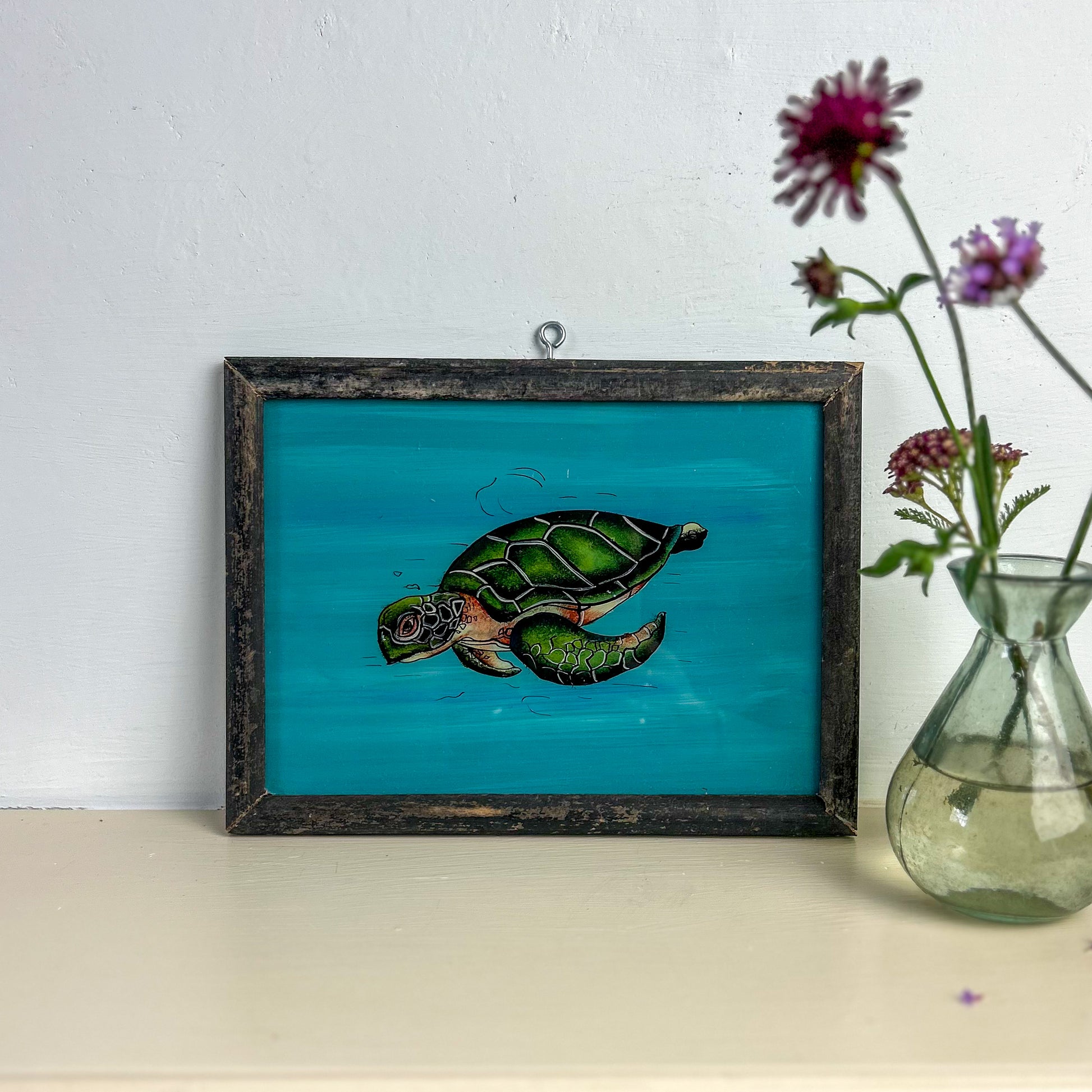 Framed artwork of a turtle on a blue background with a vase of flowers next to it. indian-glass-painting-art-reverse-turtle