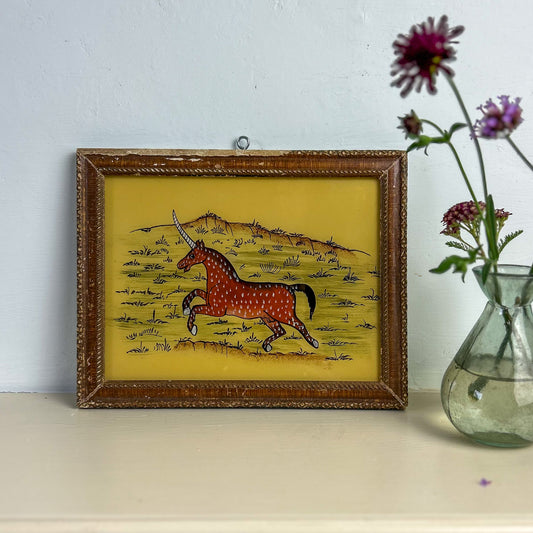 Framed artwork of a unicorn on a yellow background with a vase of flowers to the right. indian-glass-painting-art-unicorn-reverse
