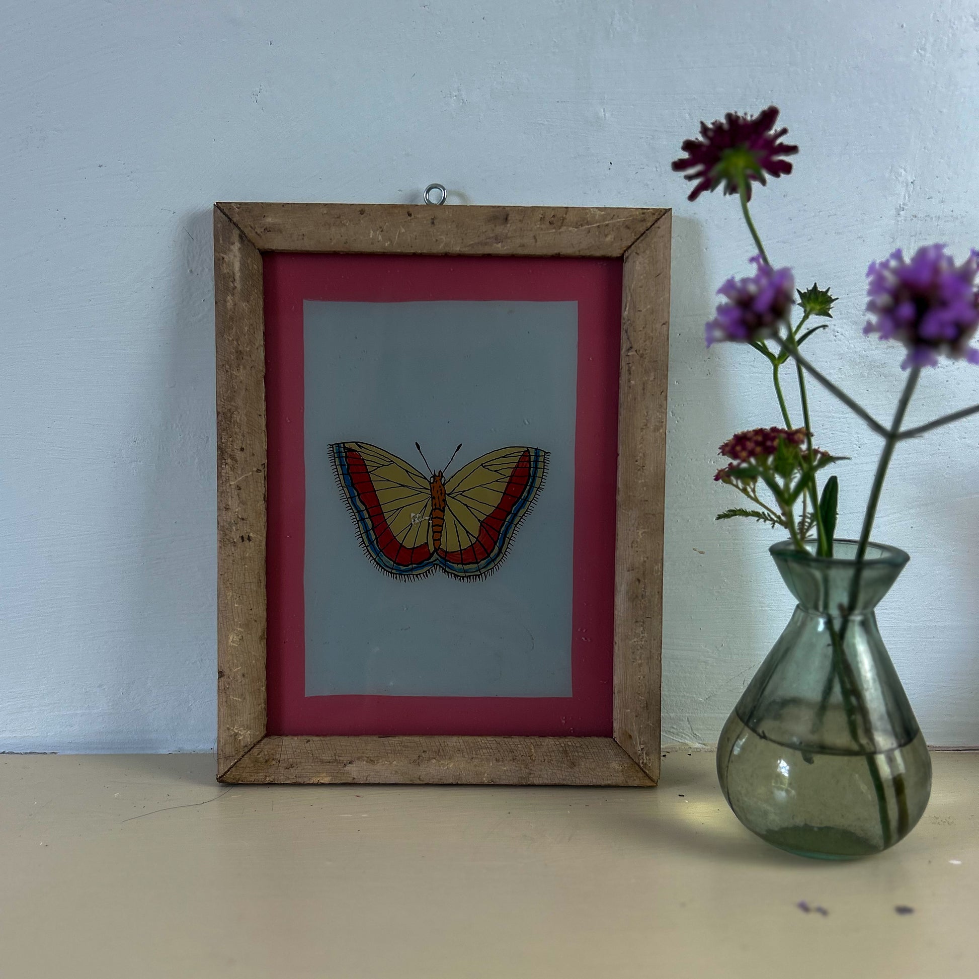 Framed butterfly artwork on a white surface with a vase of flowers. indian-glass-painting-painting-butterfly-2