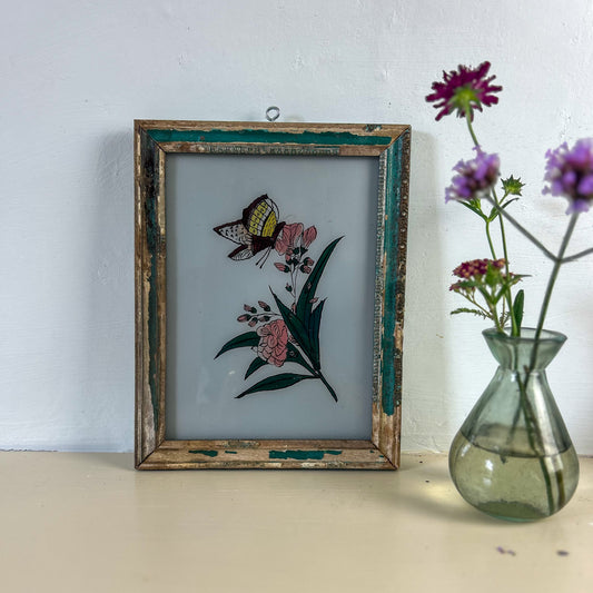 Framed embroidered artwork of a butterfly and flowers next to a clear vase with purple flowers on a light surface. indian-glass-painting-painting-butterfly-flower