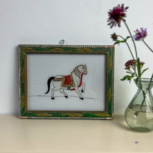 Framed artwork of a horse with a decorative border on a white surface next to a vase with flowers. indian-glass-painting-painting-horse-green-frame