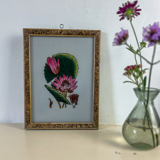 Framed botanical illustration of pink flowers and green leaves next to a vase with purple flowers on a light surface. indian-glass-painting-painting-lotus-flower