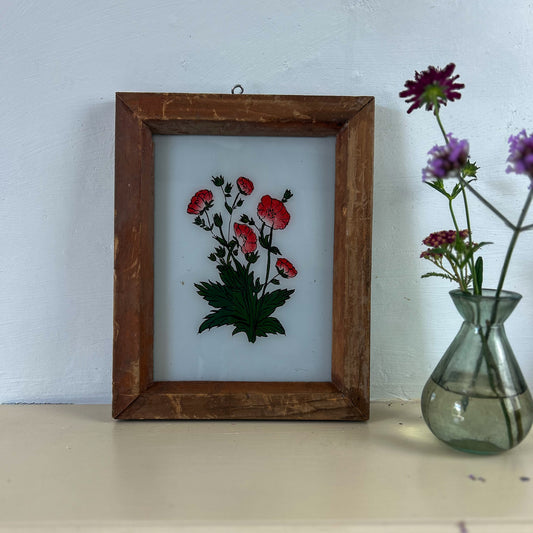 Framed embroidered artwork of flowers on a white wall with a vase of flowers beside it. indian-glass-painting-painting-red-flower-2