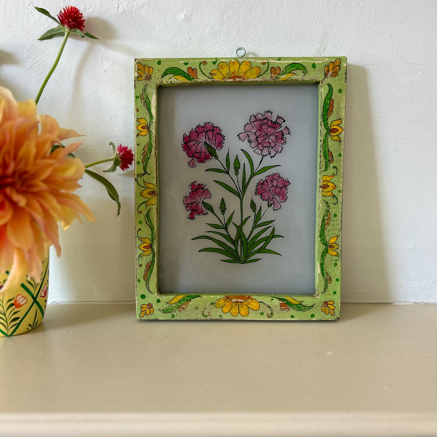 Decorative frame with floral design on a shelf with flowers. constance-and-denny-indian-reverse-glass-painting-boxed-packaging-hand-painted-artisan-art-carnations