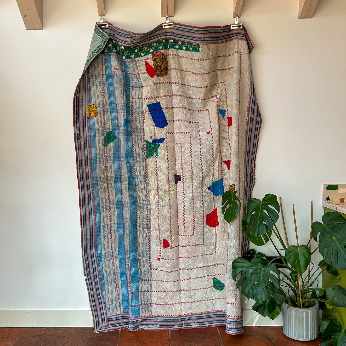 Colorful patterned skirt hanging on a rack against a white wall with a plant in the foreground. kantha-vintage-2027 - hand-made-blanket-throw-textiles-wall-hanging