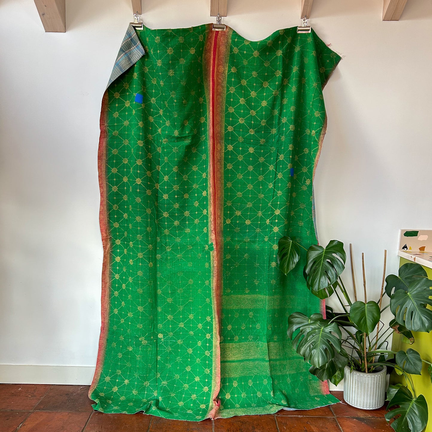 Green fabric with gold patterns draped over a wooden frame against a white wall with plants in the corner. kantha-vintage-2026 - hand-made-blanket-throw-textiles-wall-hanging