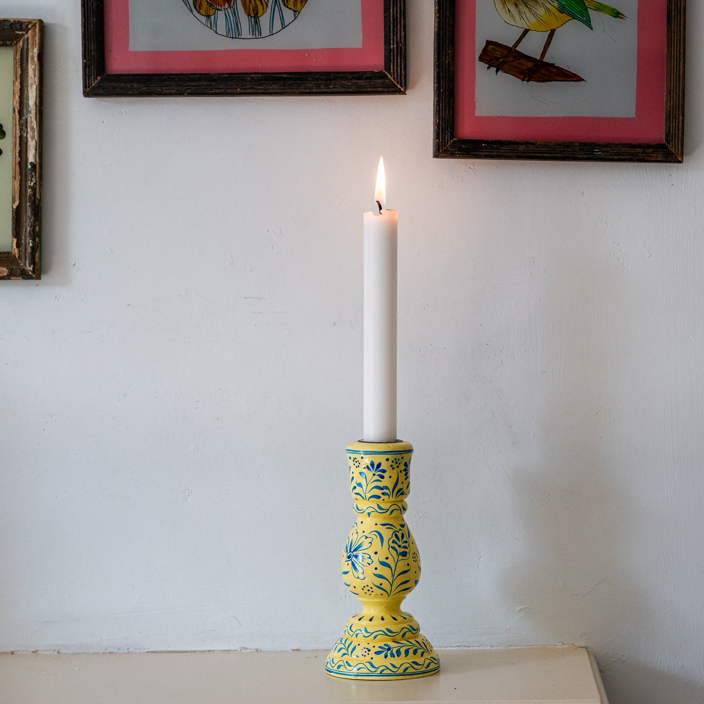 Yellow candle holder with a white candle on a white surface, framed pictures in the background. constance-and-denny-candle-stick-holder-cream-hand-painted-wooden