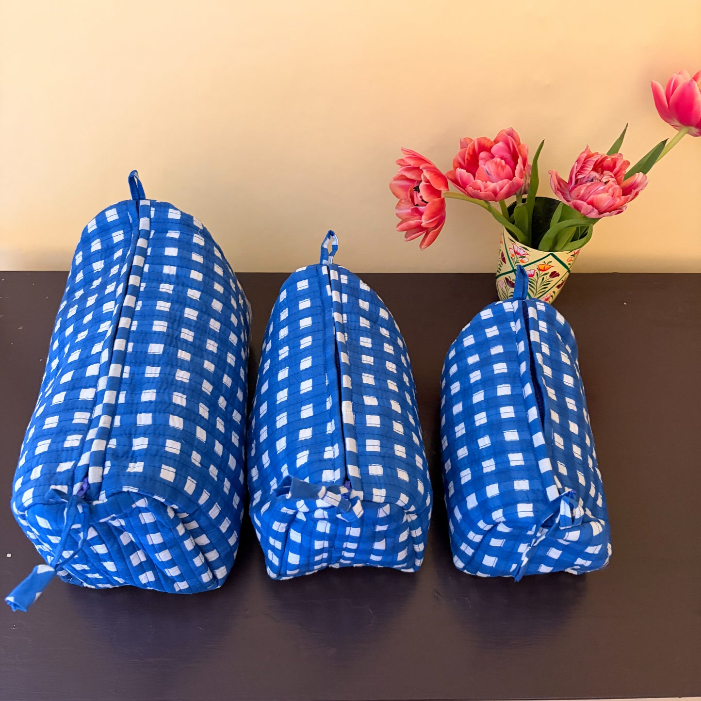 Three blue and white checkered cosmetic bags on a dark surface with pink flowers in the background.