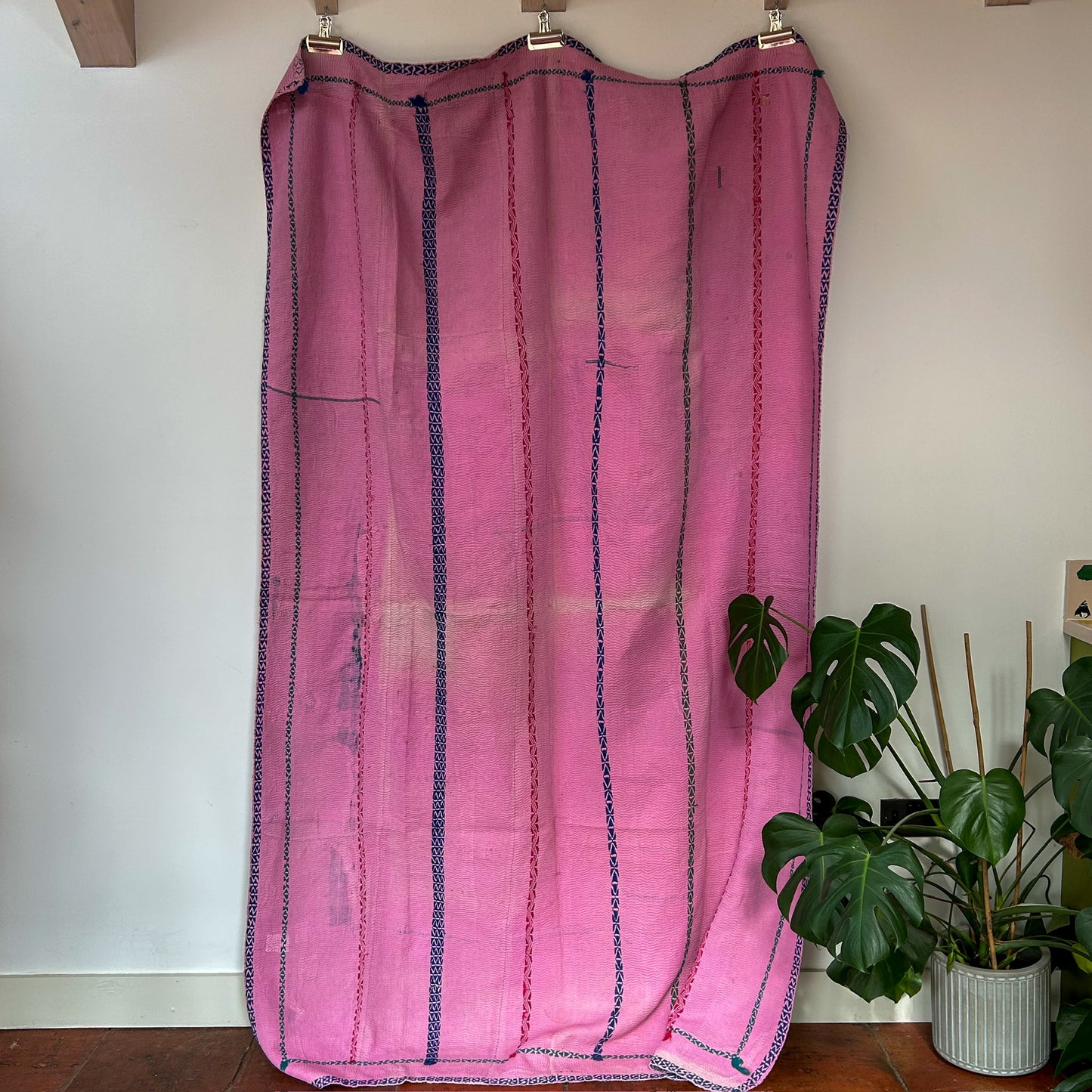 Pink fabric with blue stripes hanging on a rack against a white wall with plants in the background.