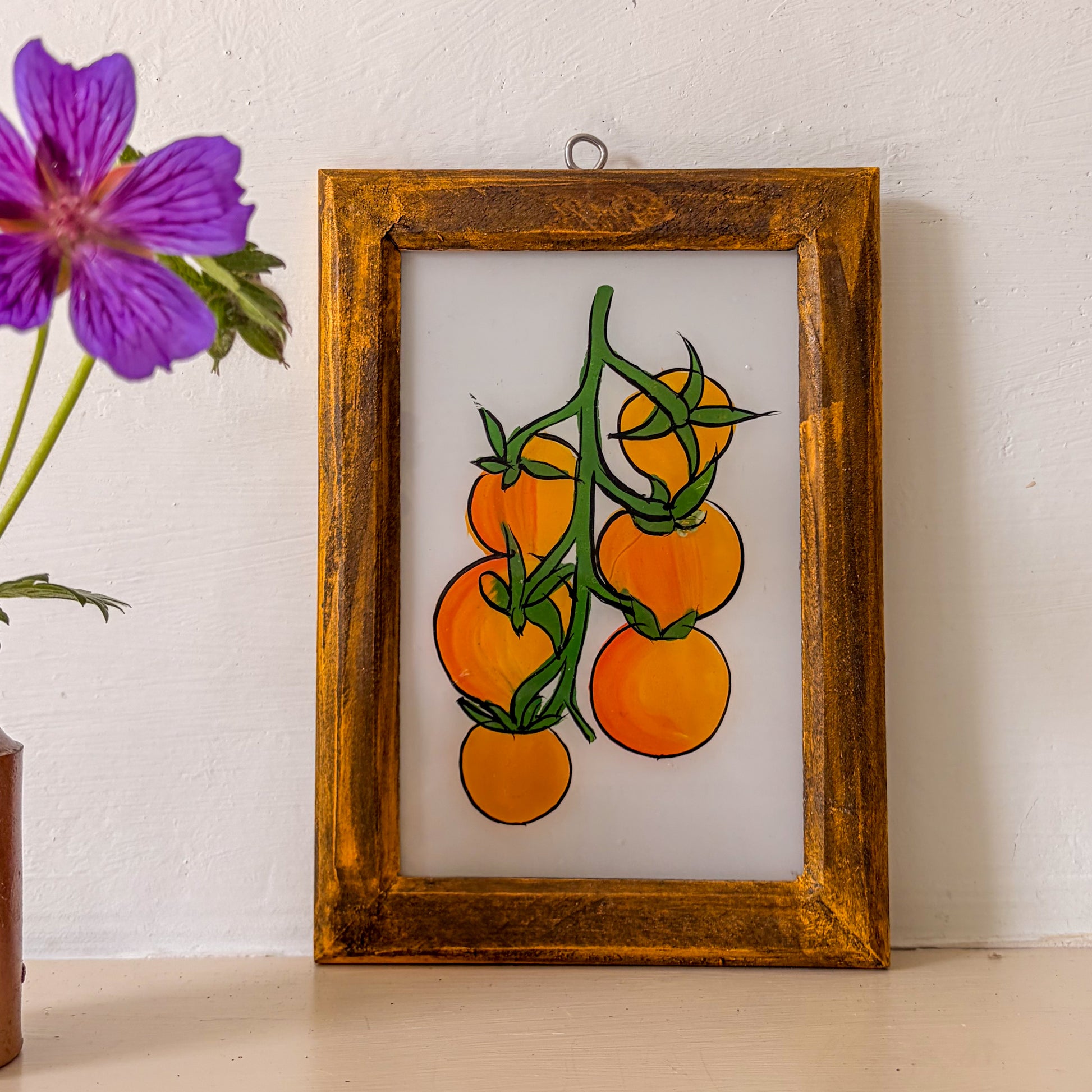 Framed artwork of orange tomatoes on a white background with a purple flower to the left.