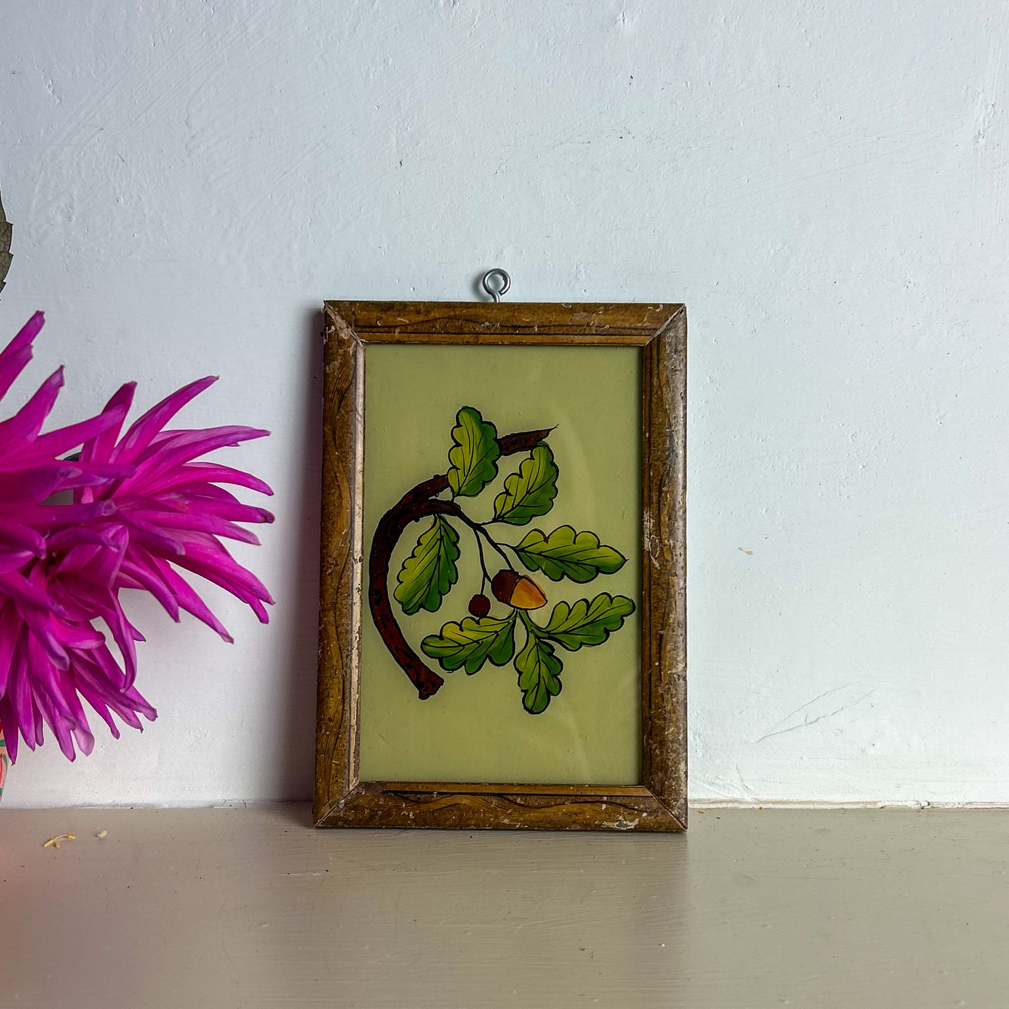 Framed artwork with green leaves and a pink flower on a white wall.