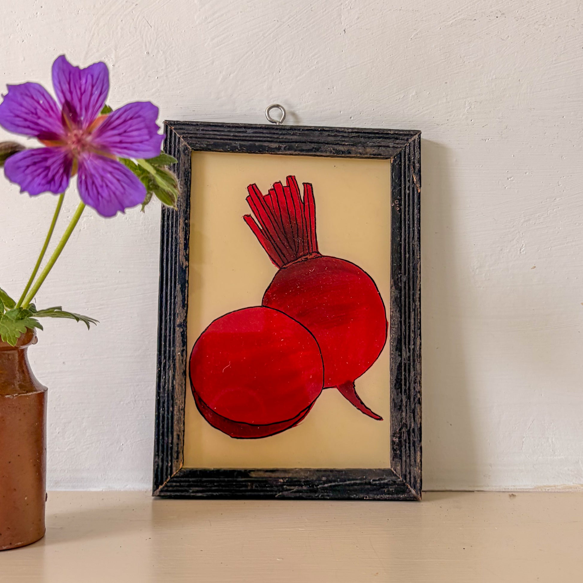 Framed stained glass artwork of two red beets on a beige background, next to a purple flower in a brown vase.