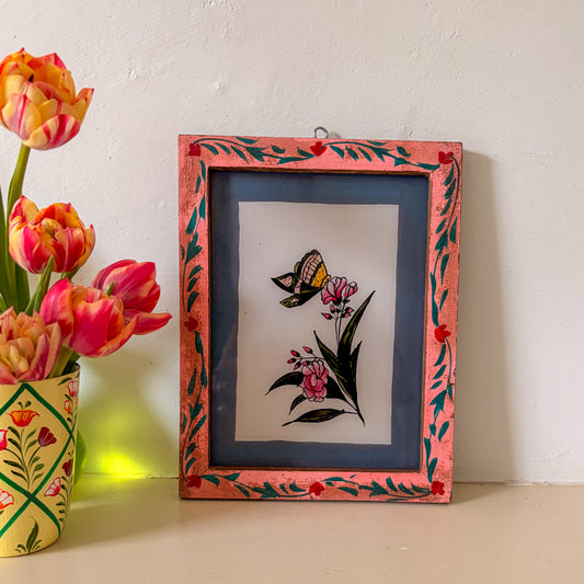 Framed artwork with a butterfly and flowers next to a vase of tulips on a white wall.