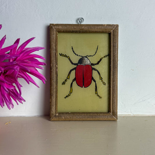 Framed artwork of a red beetle on a light background with a pink flower to the left.