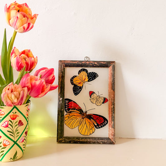 Framed artwork of butterflies next to a vase with pink tulips on a white surface.