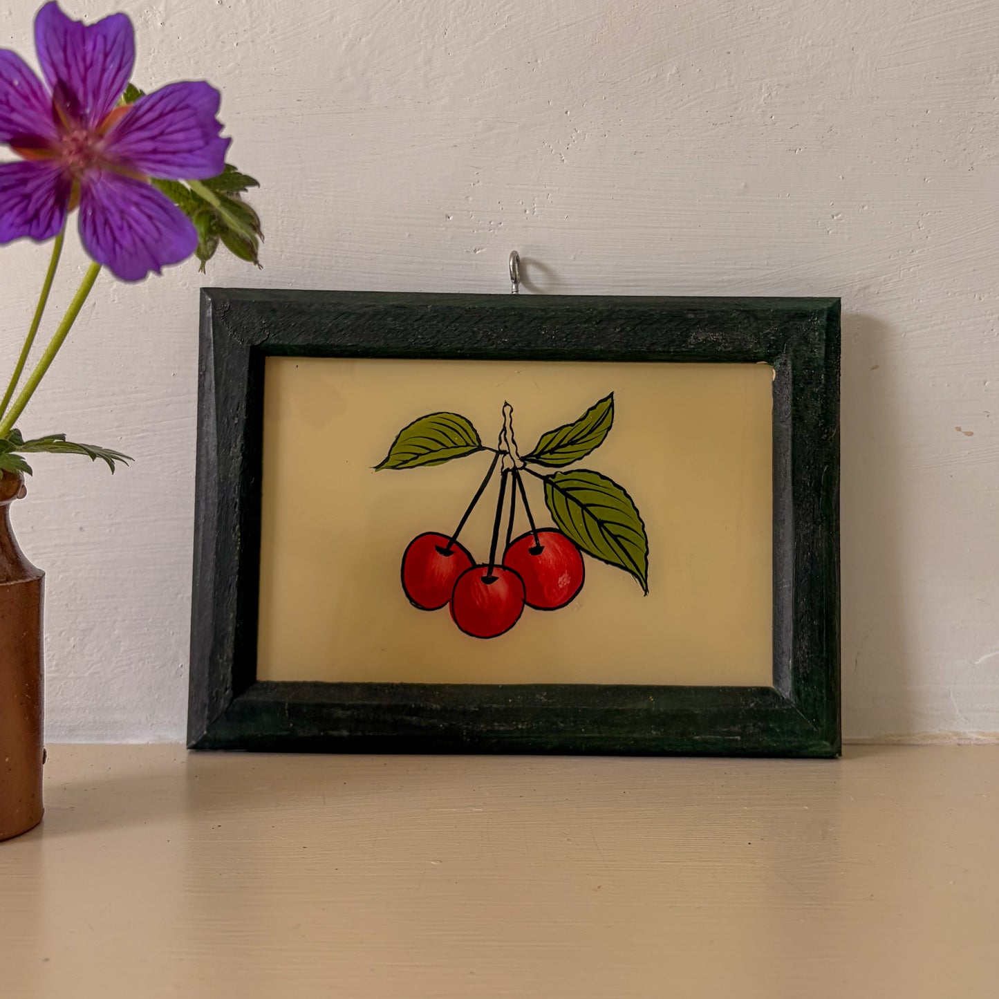 Framed artwork of cherries on a beige background with a purple flower in the corner.