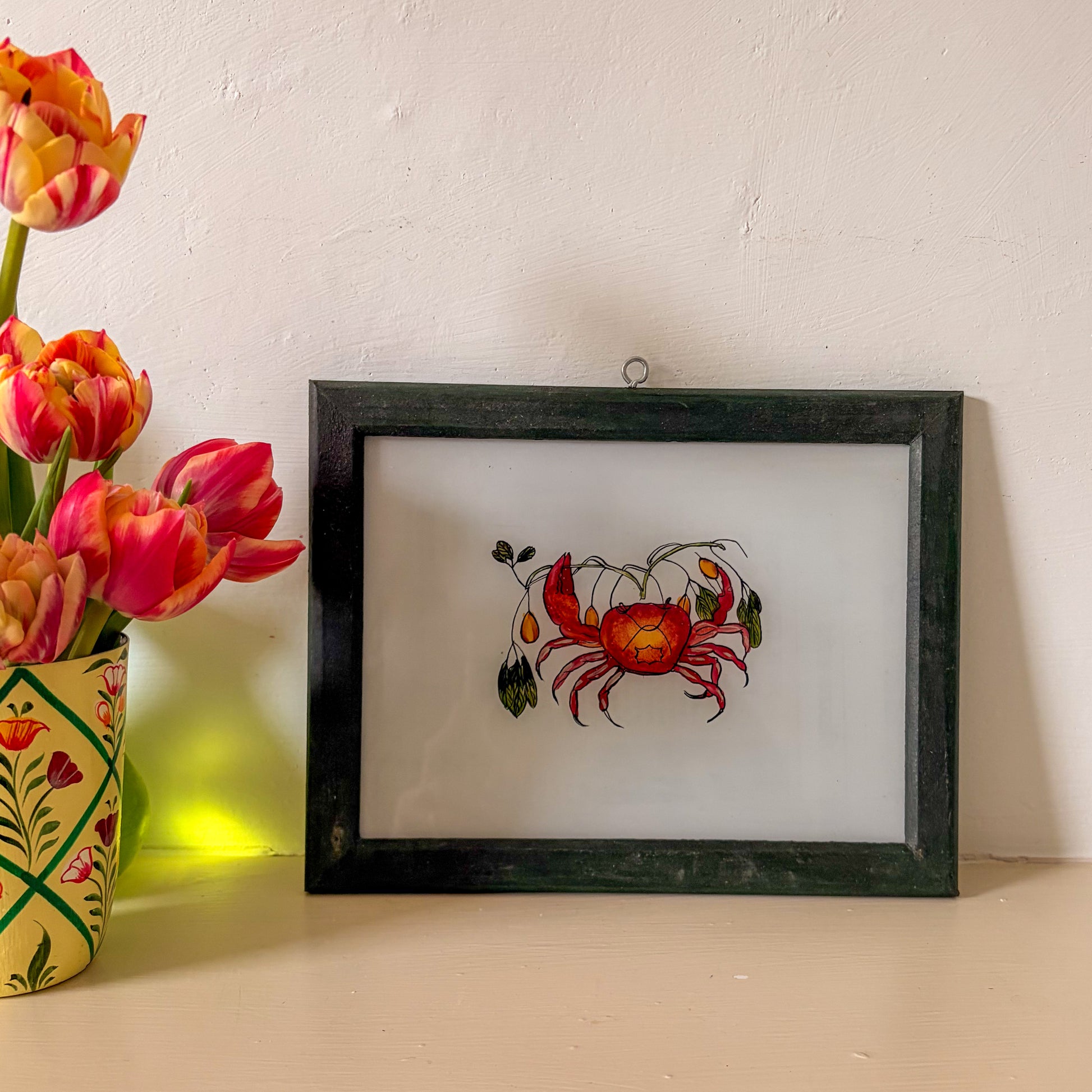 Framed artwork of a red crab with flowers on a white wall with a vase of tulips.
