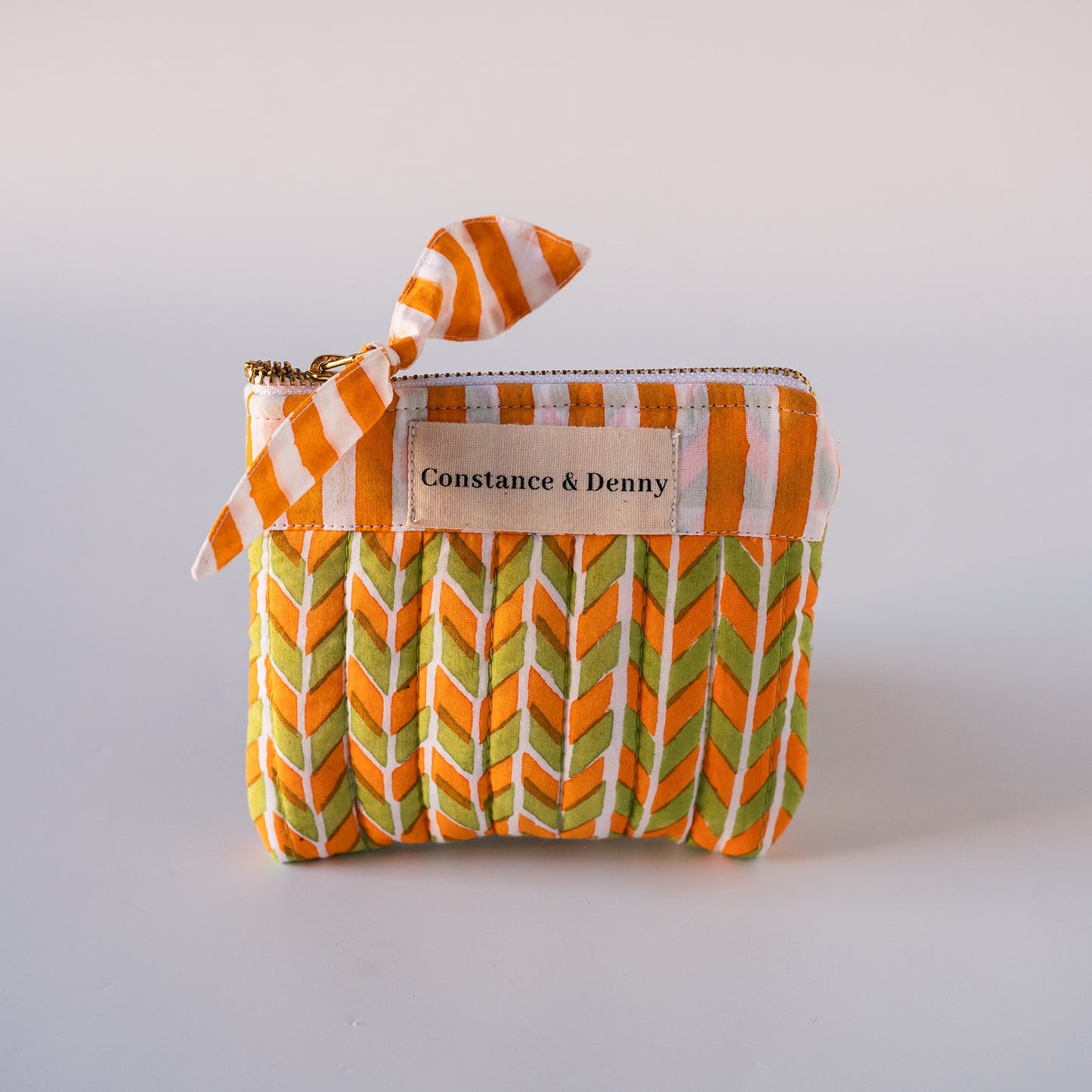 A small zippered fabric pouch with a pleated chevron pattern in orange, green, and white, featuring a matching striped ribbon tie and a beige brand label that reads 'Constance & Denny'.