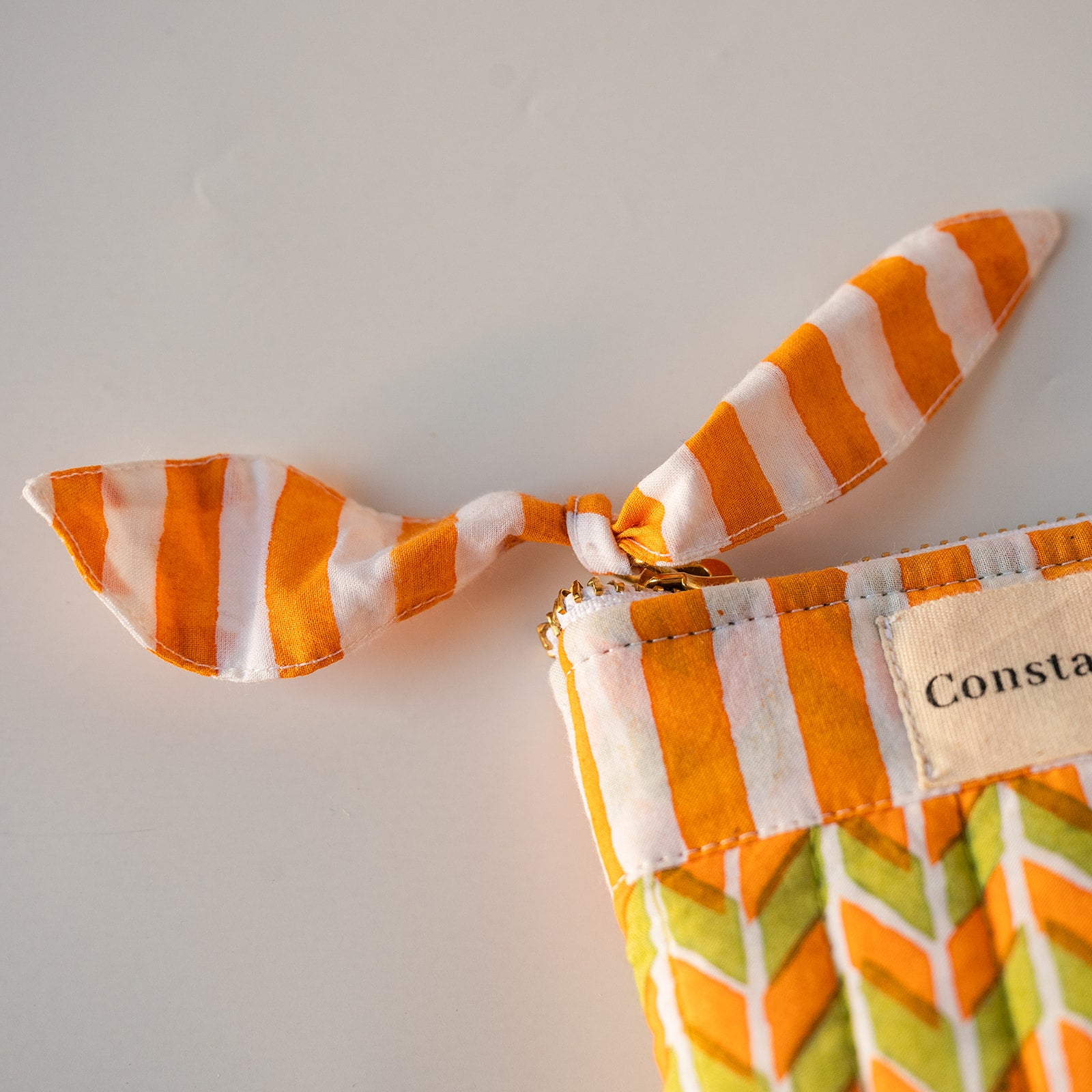 Close-up of a fabric zip pouch with orange and white striped ribbon ties, a cream fabric label reading 'Consta', and a green and orange chevron patterned fabric body, set against a plain light background.