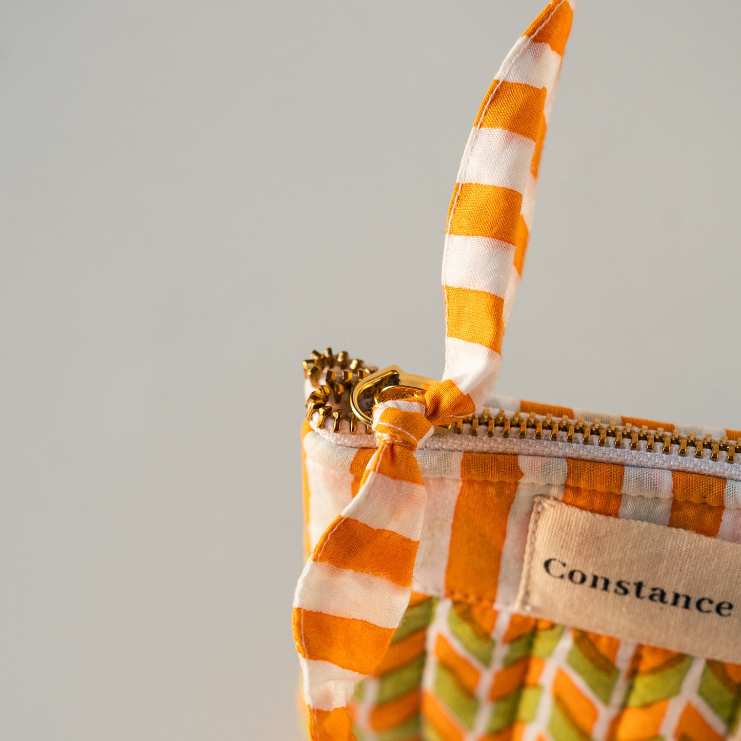 Close-up of a Constance brand zippered pouch featuring a vibrant orange and white striped fabric handle and a beige canvas label, set against a neutral light gray background.