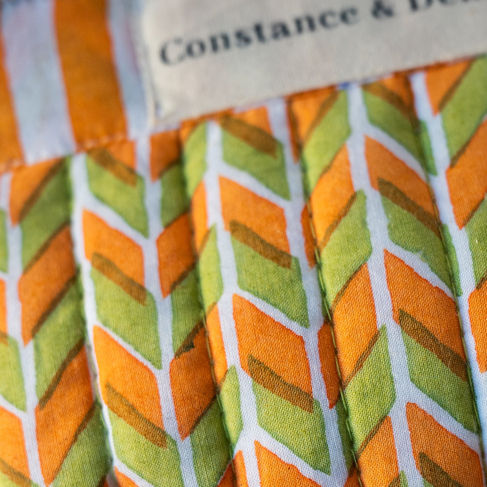 Close-up of a patterned fabric item with orange, green, and white chevron-like stripes, plus part of a beige fabric label reading 'Constance & De.
