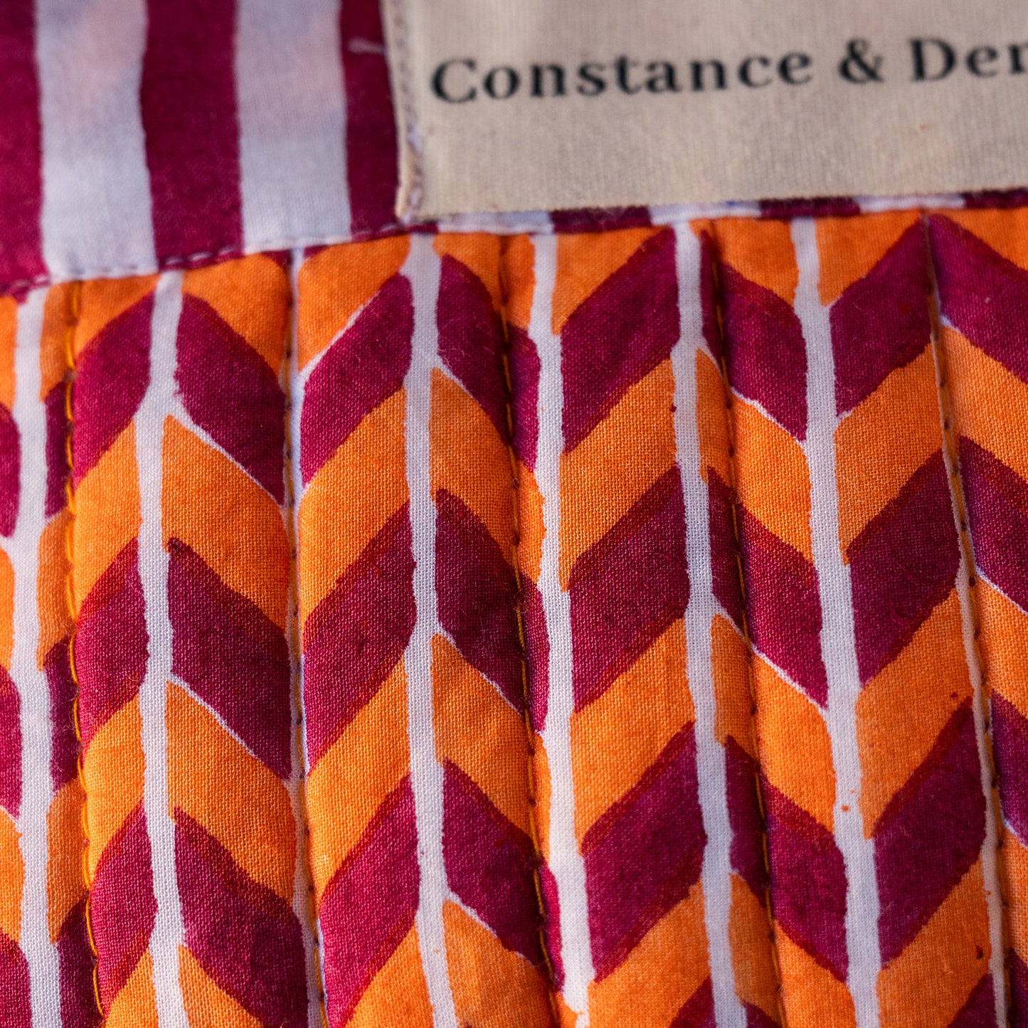 Close-up of a pleated fabric item with a bold chevron pattern in orange, burgundy, and white, featuring a stitched label that reads 'Constance & Der'.