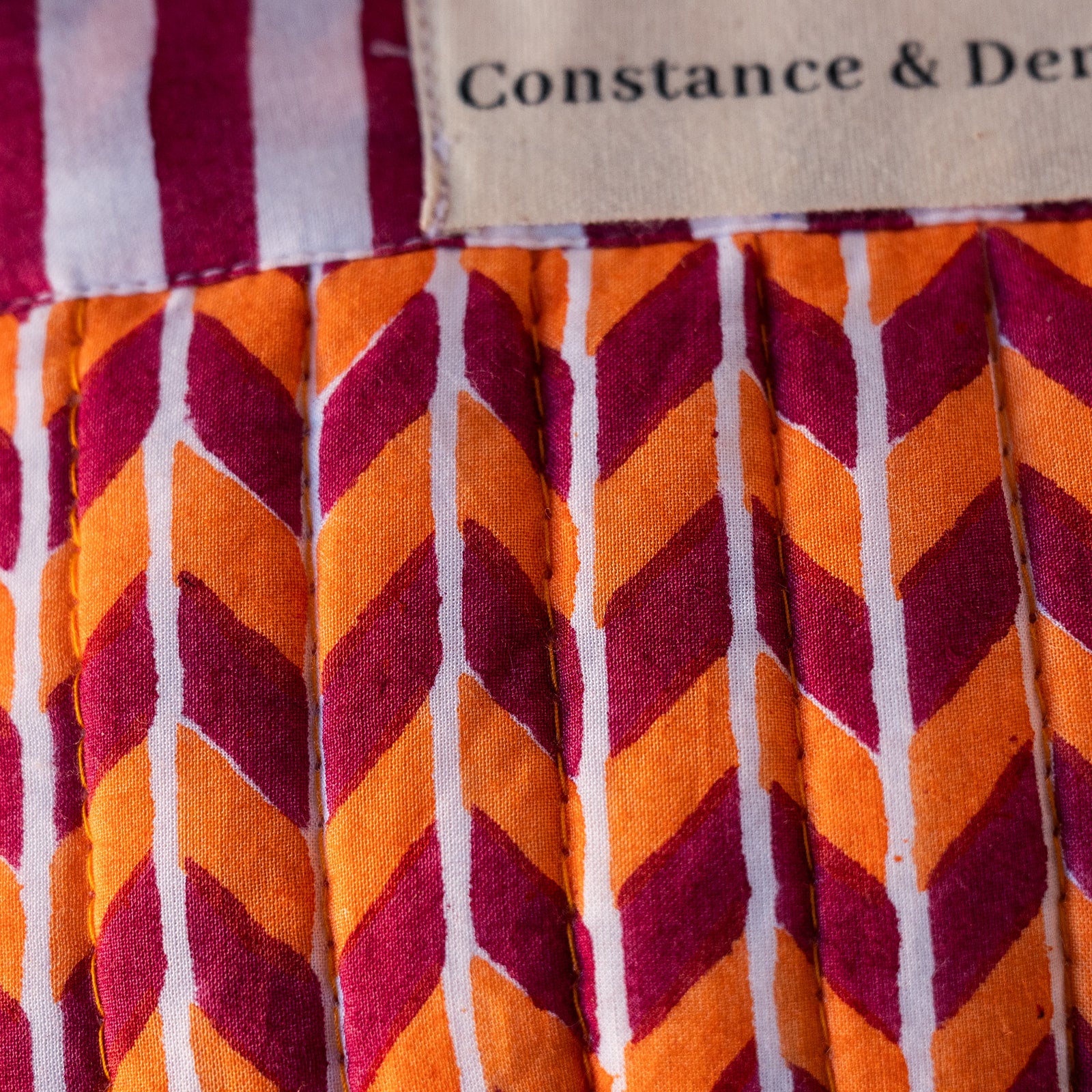Close-up of a pleated fabric item with a bold chevron pattern in orange, burgundy, and white, featuring a stitched label that reads 'Constance & Der'.