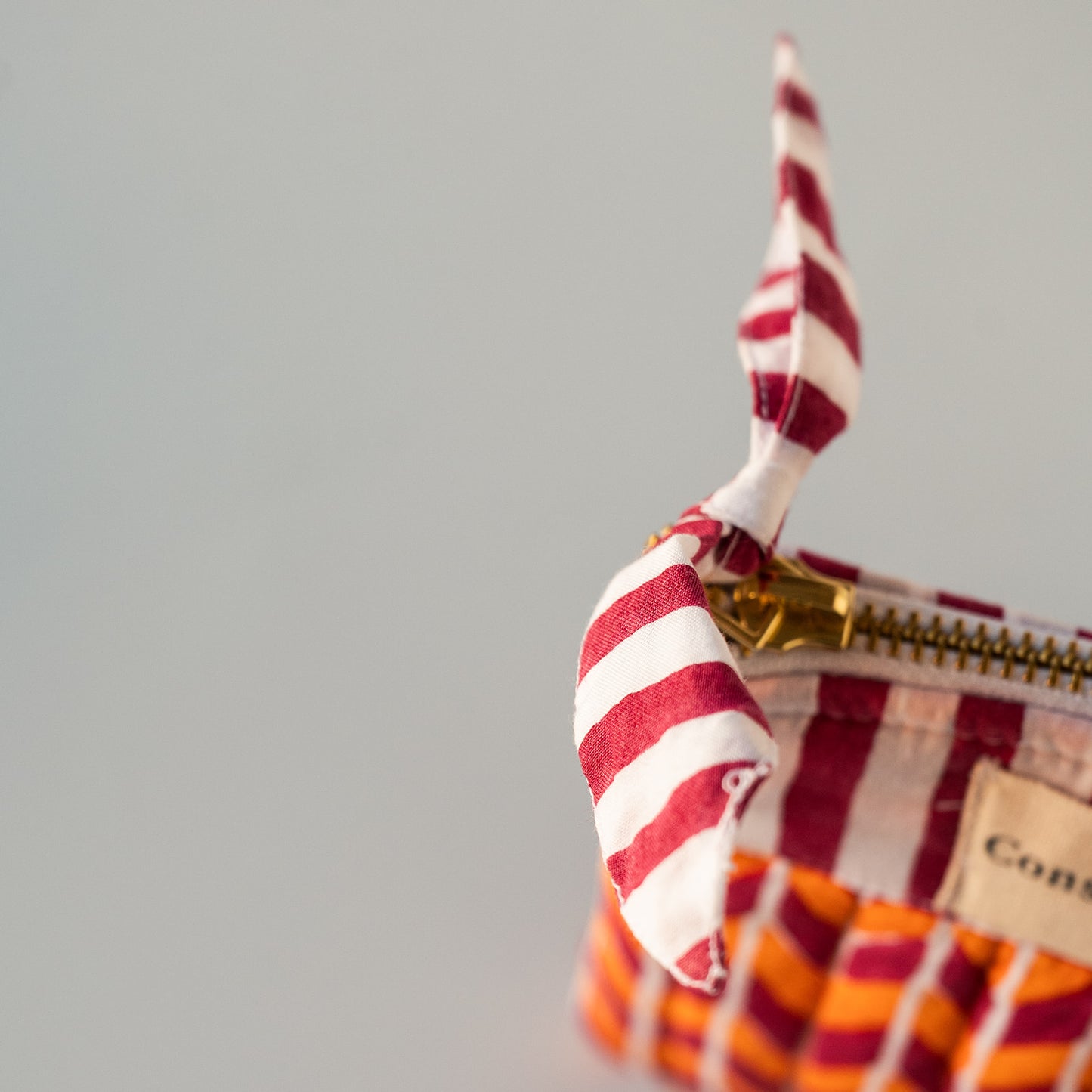Close-up of a Constance brand zippered pouch featuring a vibrant orange and white striped fabric handle and a beige canvas label, set against a neutral light gray background.
