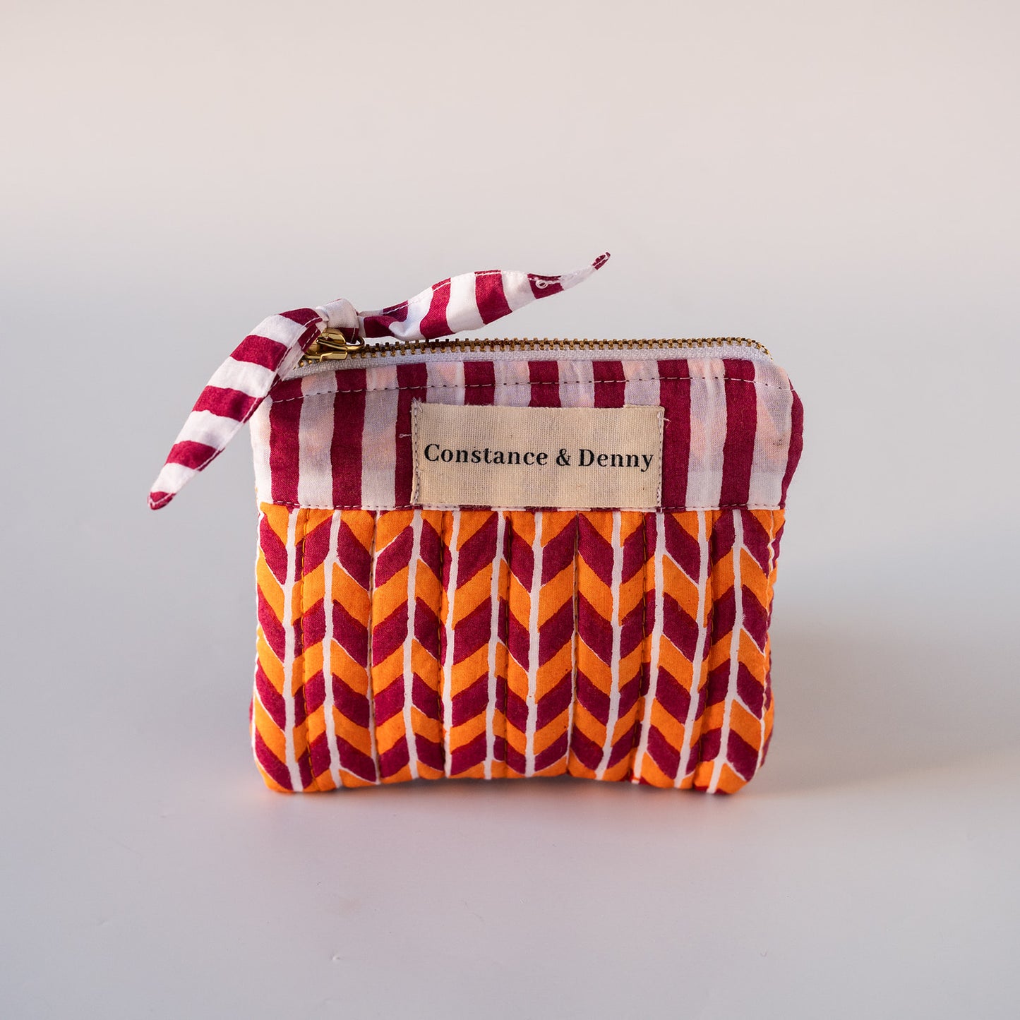 A small zippered fabric pouch with a pleated chevron pattern in orange, green, and white, featuring a matching striped ribbon tie and a beige brand label that reads 'Constance & Denny'.
