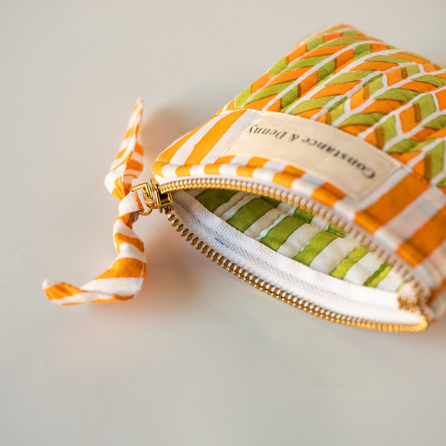 A close-up of a small zippered pouch with a woven orange and green patterned exterior, a cream fabric label reading 'Constance & Deny,' and a gold zipper. The pouch is shown open on a clean white background.