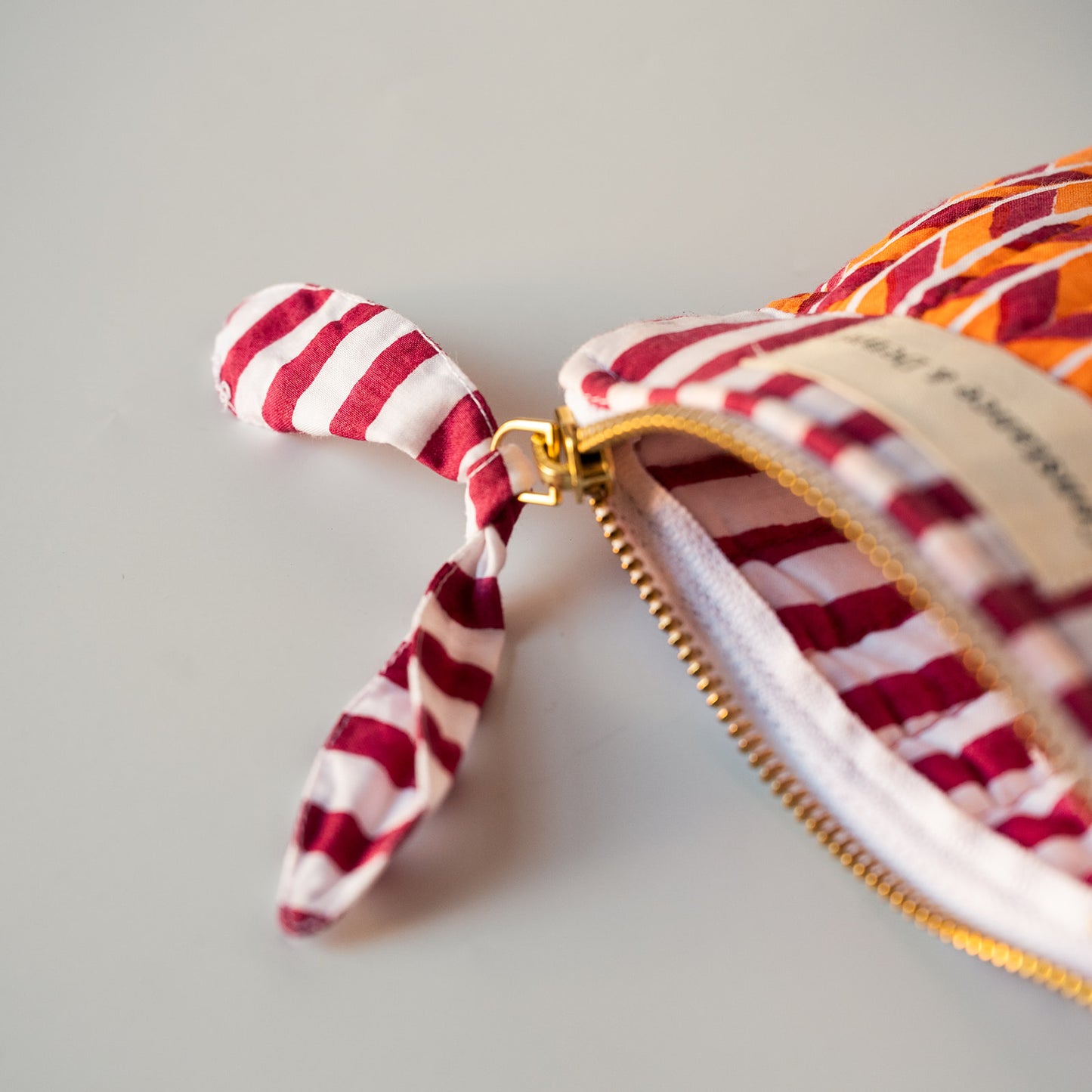 A close-up of a fabric zippered pouch with a small hanging ribbon pull. The pouch has a red and white striped pattern, a gold zipper, and a cream fabric label.