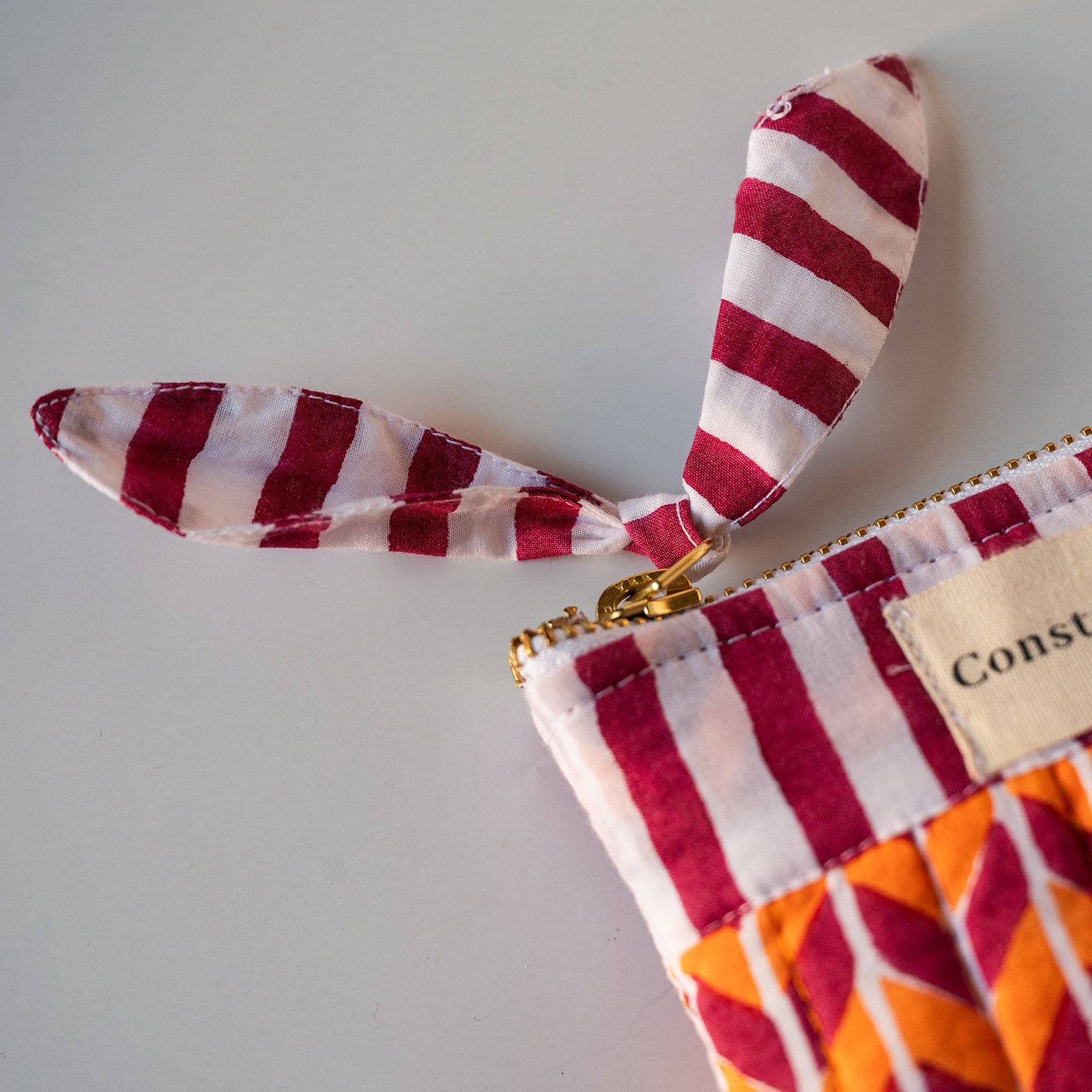 A close-up of a fabric zip pouch with bunny ears, featuring a red and white striped pattern, a gold zipper, and an orange and magenta geometric patterned section. A beige fabric label with the partial text 'Const' is visible.