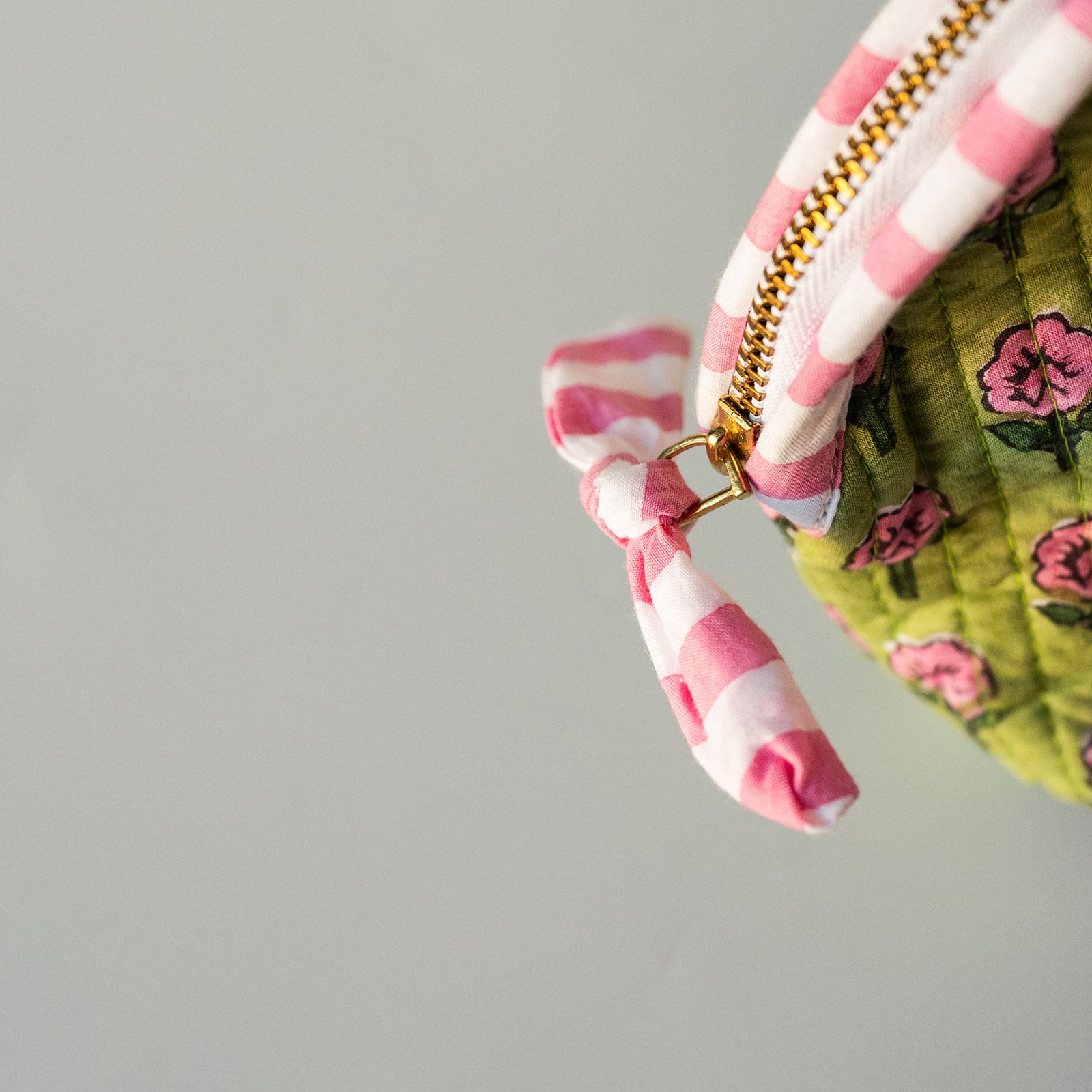 Close-up of a fabric zipper pull and part of a quilted pouch. The zipper pull is pink and white striped with a small gold metal ring, next to a green floral quilted fabric.